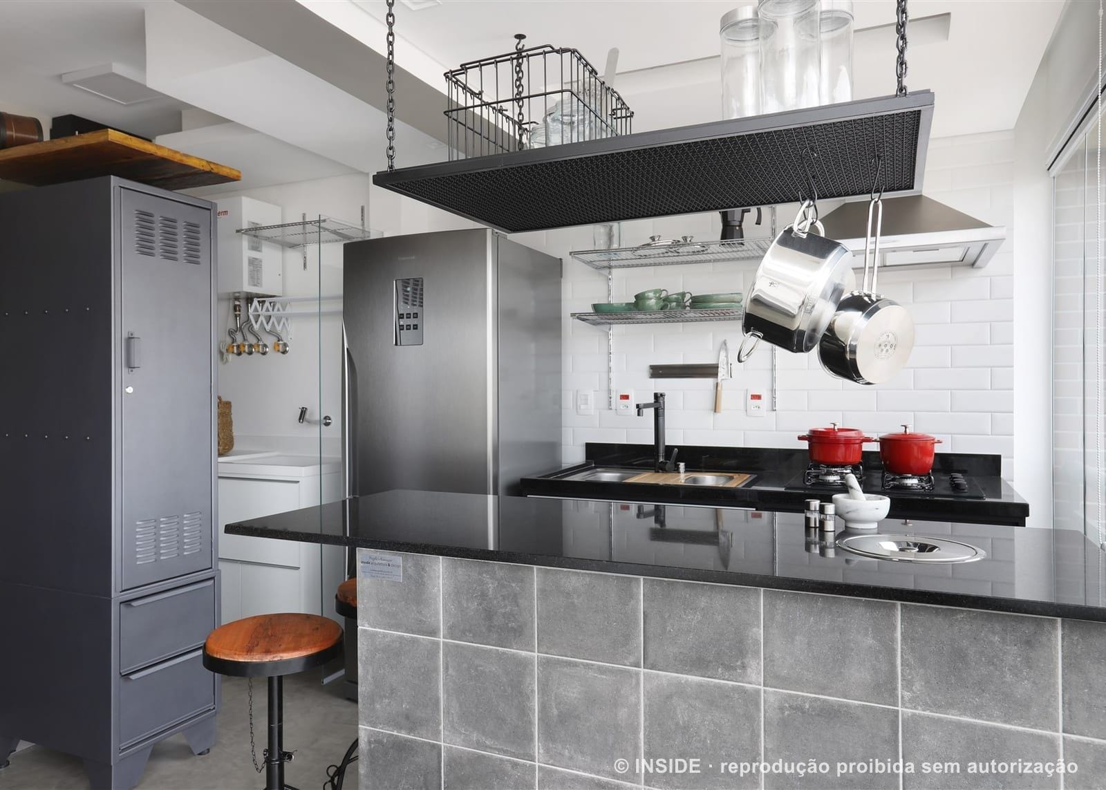 Cozinha aberta com ilha revestida de cimento, bancada de granito preto, cooktop a gás, cuba embutida e estrutura suspensa de panelas ancorada no teto, projeto INSIDE