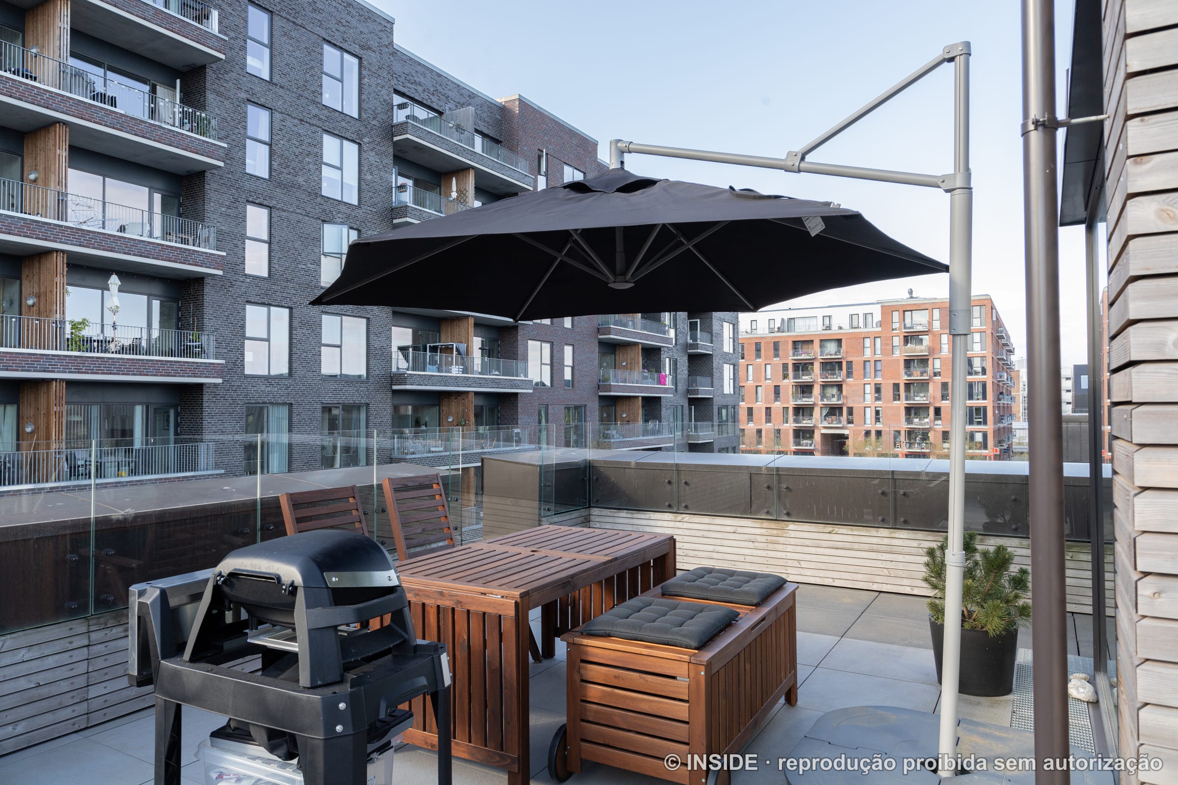 Terraço externo com mesa e bancos de madeira, ombrelone preto, churrasqueira e guarda-corpo de vidro, projeto INSIDE