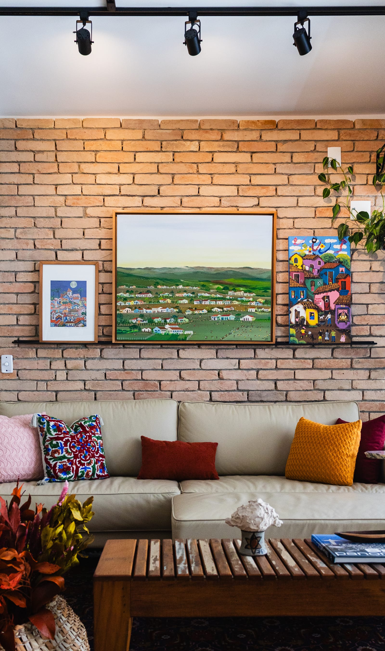Sala com parede de tijolo aparente e trio de quadros naïf sobre o sofá caramelo, mesa de demolição à frente, projeto INSIDE