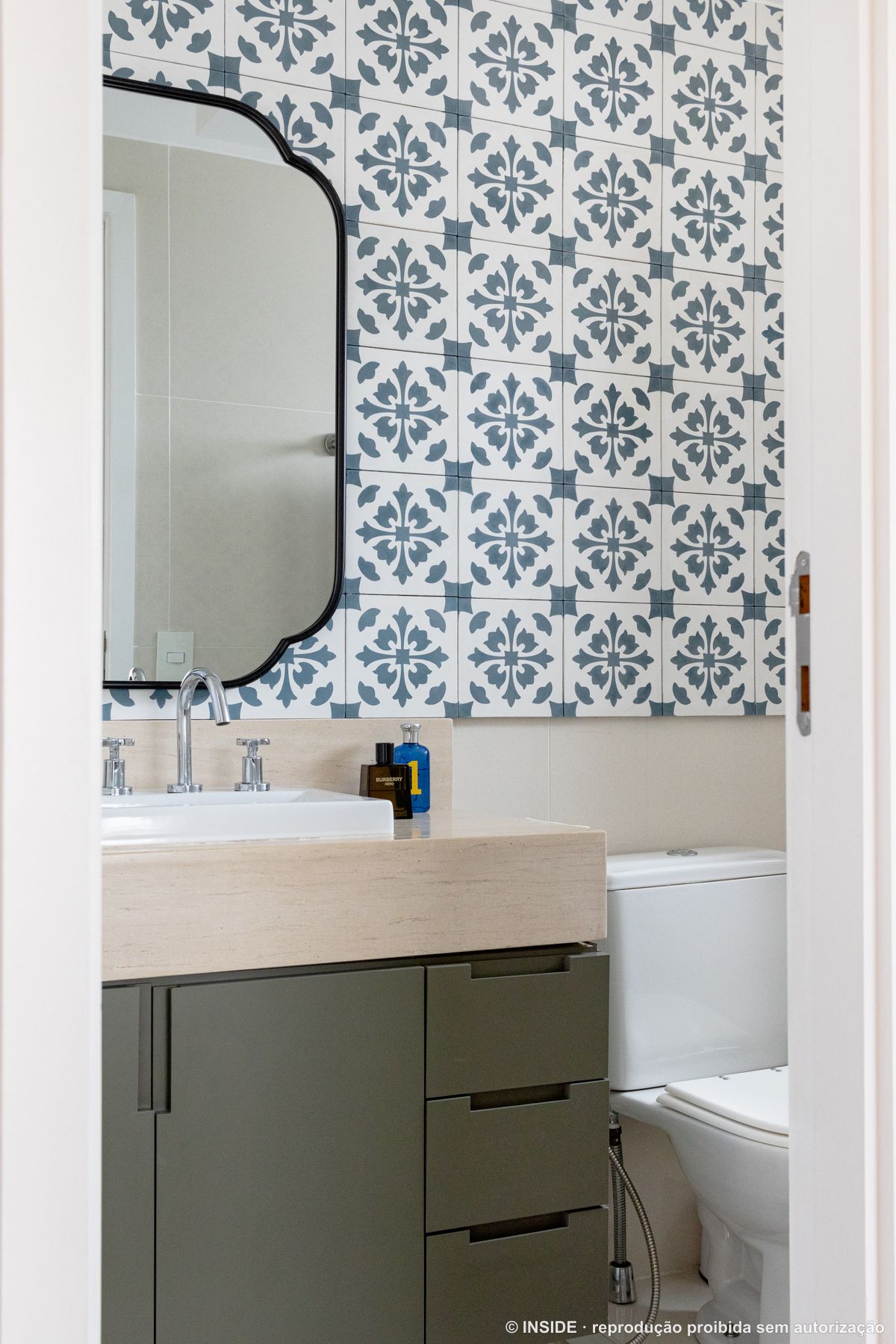 Banheiro da suíte com parede de ladrilho hidráulico azul e branco, espelho de moldura preta recortada, bancada de travertino e marcenaria verde-oliva, projeto INSIDE