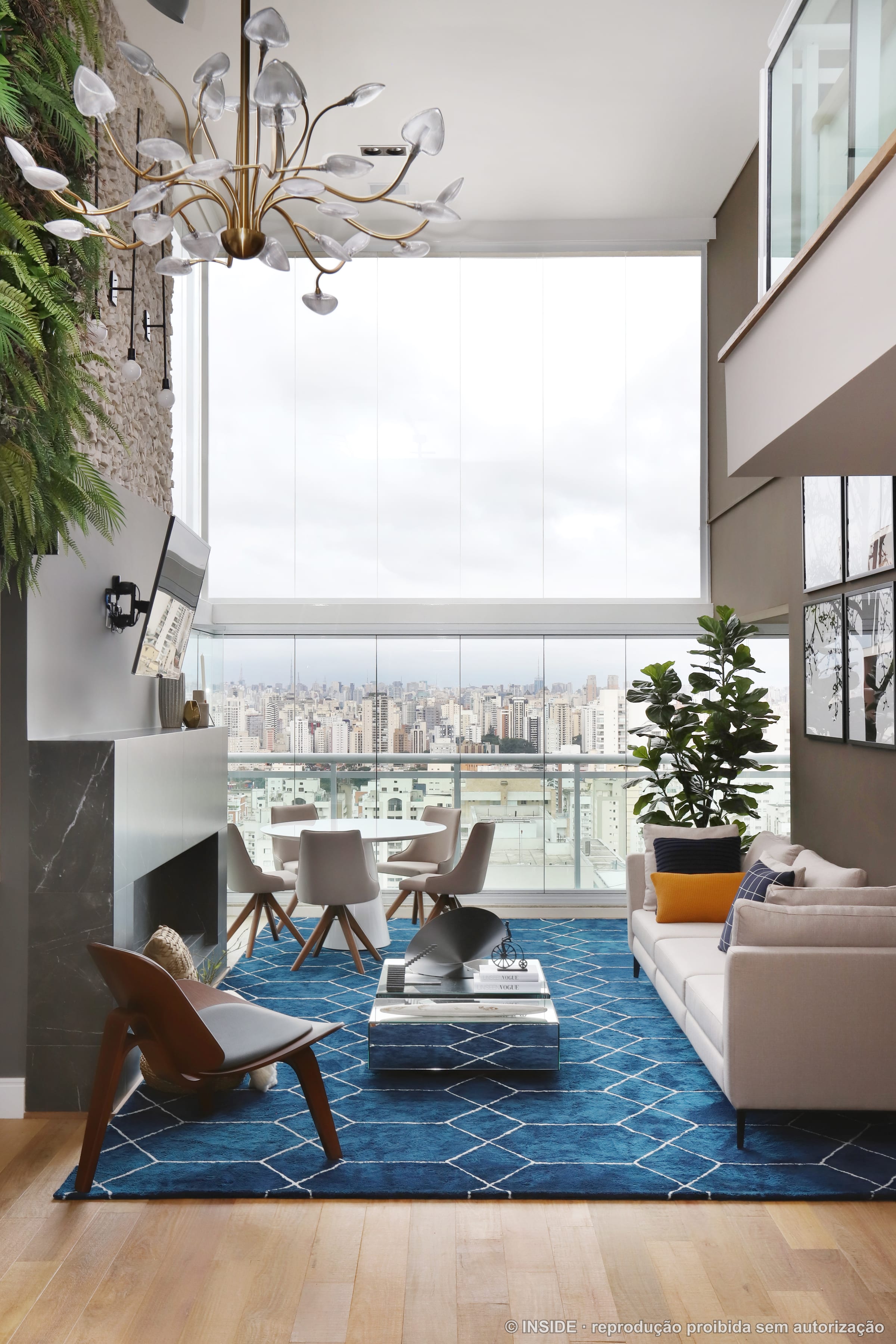 Sala de estar ampla de pé-direito duplo com lustre de hastes, poltrona de design, tapete azul, painel de TV e vista da cidade, projeto INSIDE