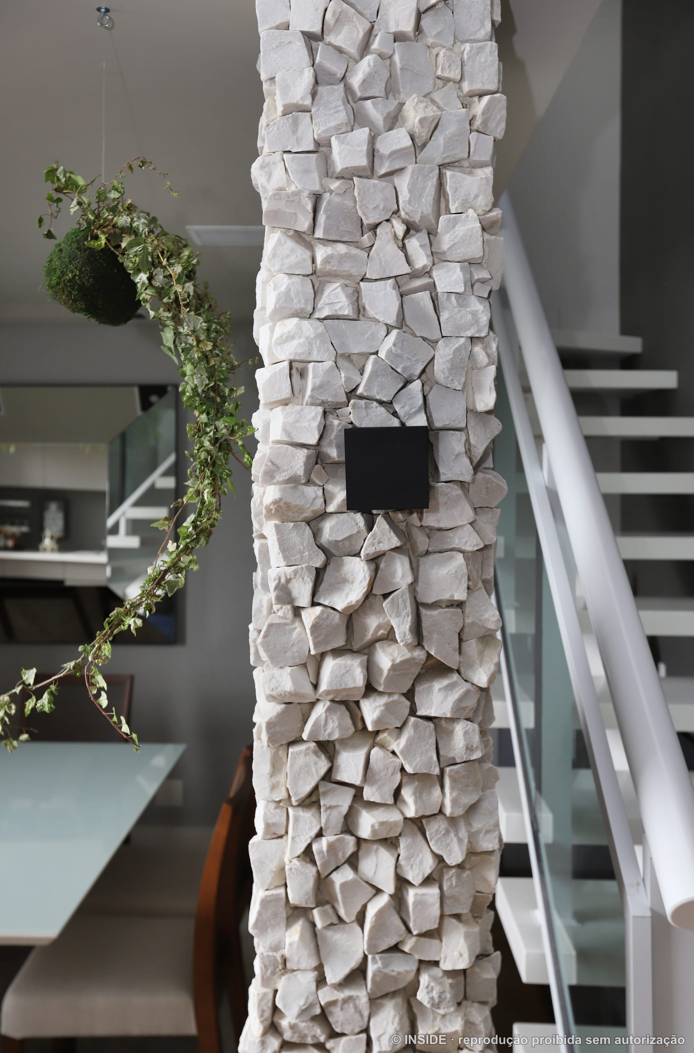 Detalhe da sala com coluna revestida em pedra bruta, escada metálica branca e trepadeira verde descendo pelo pé-direito duplo, projeto INSIDE