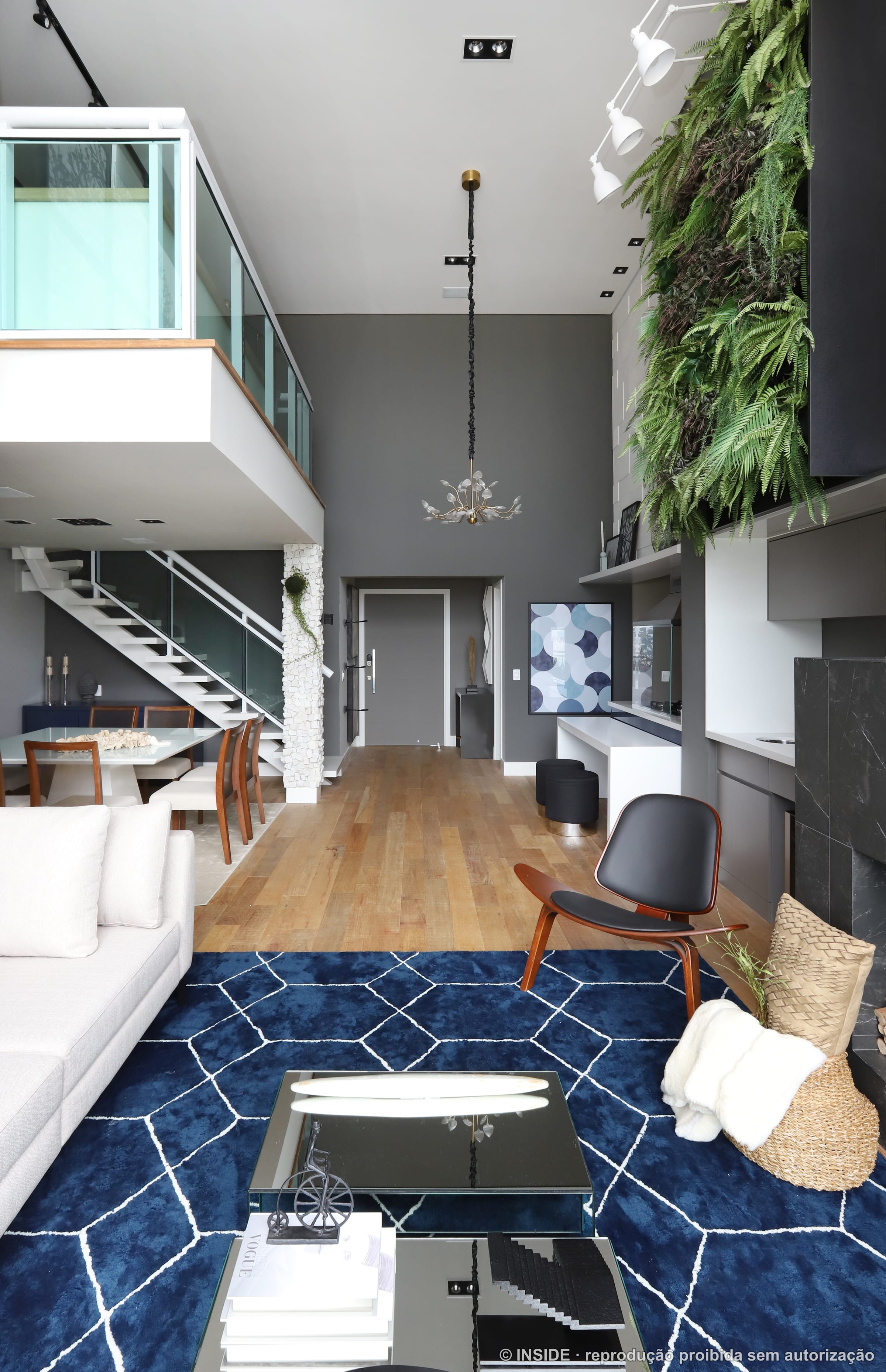 Sala de estar com parede de vegetação viva, escada metálica, sofá neutro, poltrona de design e tapete azul geométrico, projeto INSIDE