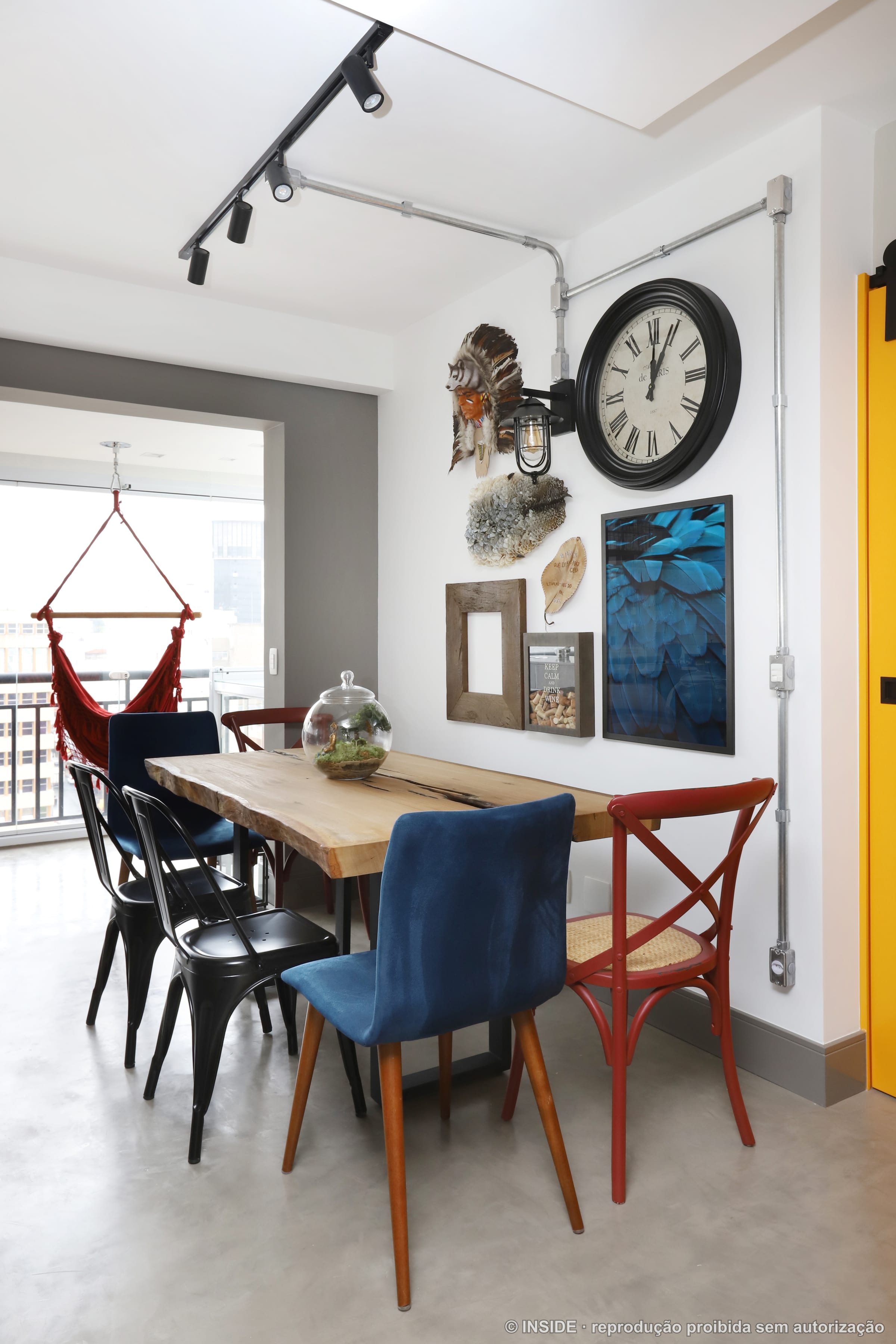 Sala de jantar com mesa de madeira, cadeiras azuis, parede de quadros e relógio, com rede ao fundo, projeto INSIDE
